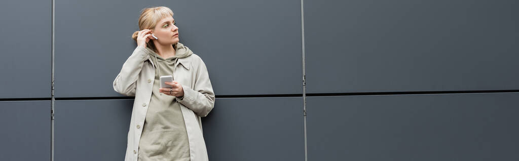 elegante giovane donna con frangia in piedi in abiti alla moda e auricolari wireless durante l'utilizzo di smartphone vicino grigio edificio moderno sulla strada urbana, cappotto con cappuccio, guardando altrove, banner  - Foto, immagini