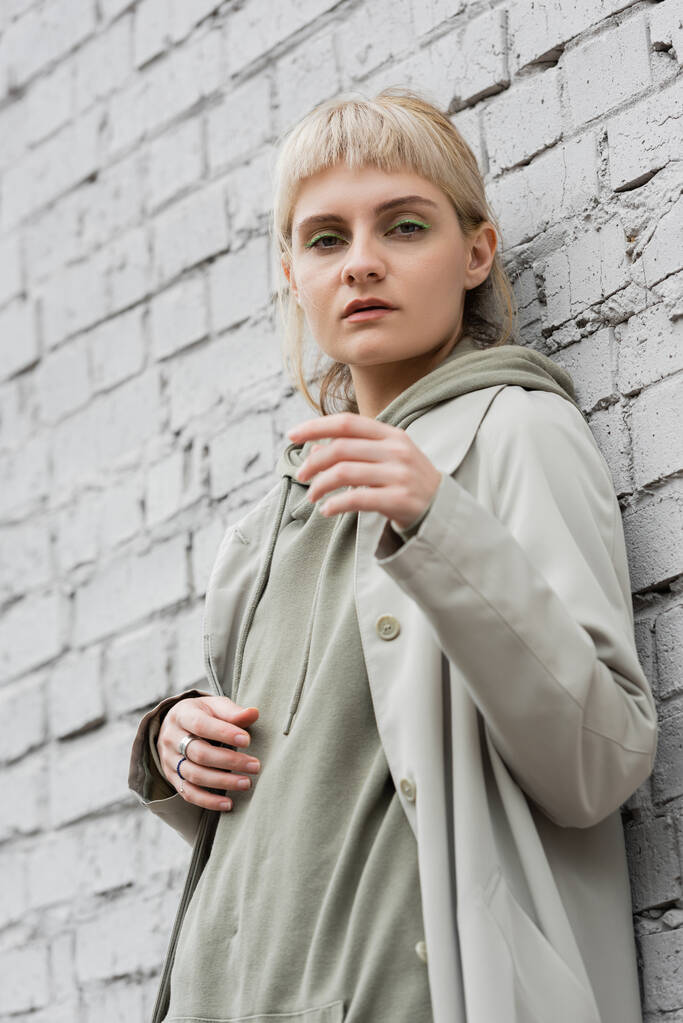 mladá žena s make-upem, blond vlasy, ofina, ve stylovém oblečení stojí v blízkosti šedé cihlové zdi moderní budovy a při pohledu na kameru při pózování venku, kabát, módní trend  - Fotografie, Obrázek