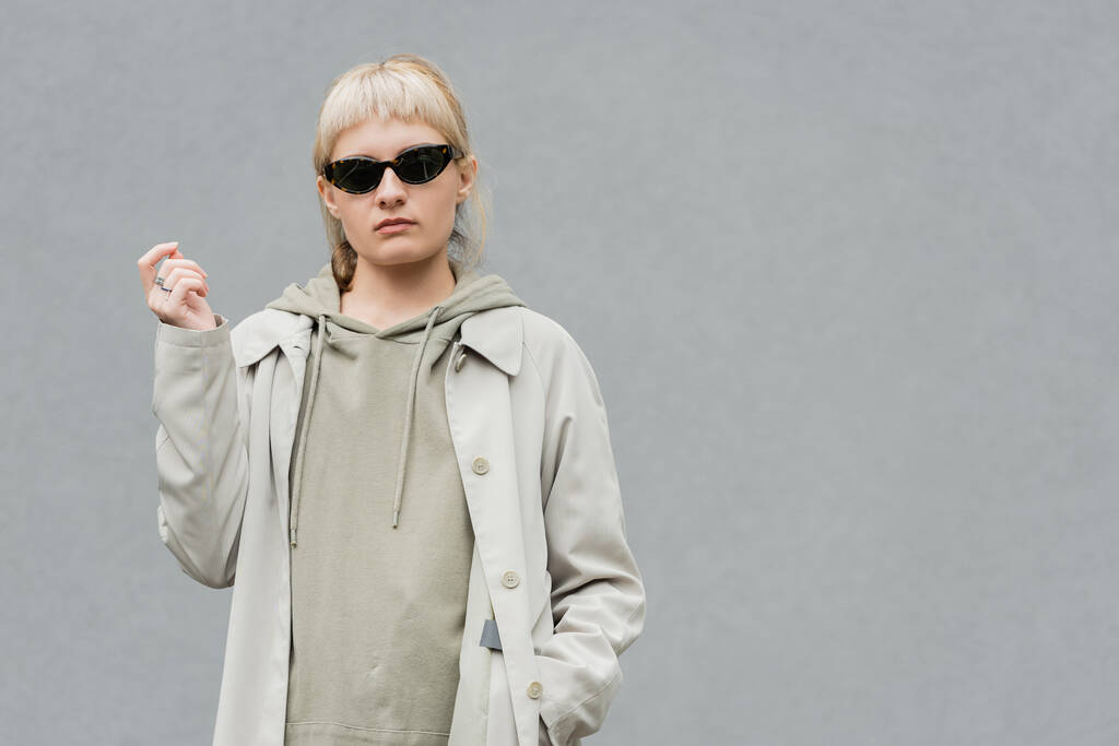 stylish and young woman with bangs and blonde hair standing in trendy sunglasses and comfortable clothes while looking at camera and posing with hand in pocket isolated on grey background in studio  - Photo, Image