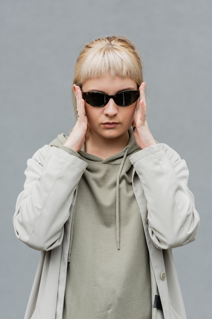 stylish and young woman with bangs and blonde hair standing in trendy sunglasses and comfortable clothes while looking at camera isolated on grey background in studio, hoodie  - Photo, Image
