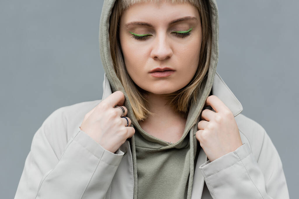 stylish and young woman with bangs, green eye shadows and blonde hair standing with hood on head and comfortable clothes while looking down isolated on grey background in studio, hoodie  - Photo, Image