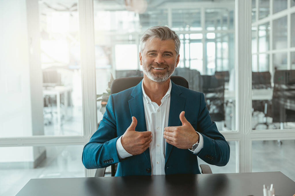 Улыбающийся взрослый бизнесмен, показывающий поднятые пальцы, сидя в офисе. Высокое качество фото - Фото, изображение