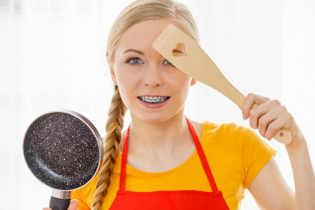 Jovem alegre usando avental segurando pequena panela de cozinha e espátula de madeira prestes a cozinhar na cozinha. - Foto, Imagem