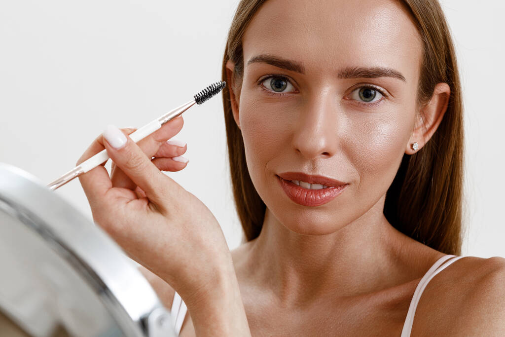 Young woman combing her eyebrows with brow brush over white studio background. Fnishing makeup - Photo, Image