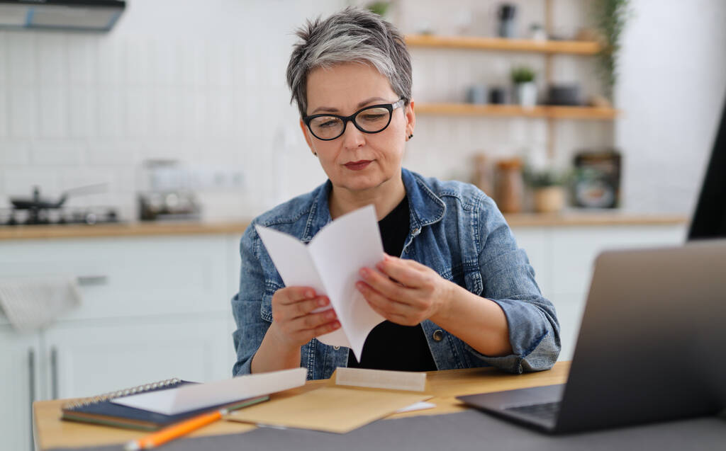 Een serieuze vrouw met een bril kijkt naar de post en bankafschriften aan de tafel naast de laptop. - Foto, afbeelding