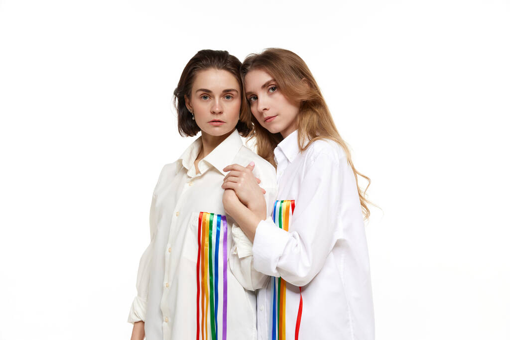2人の若い女性の肖像画、ルブトの愛を象徴する虹のラインと白いシャツのカップル。モデルは白を背景にポーズ。lgbtコミュニティ、支援、愛、人権、プライド月間の概念 - 写真・画像
