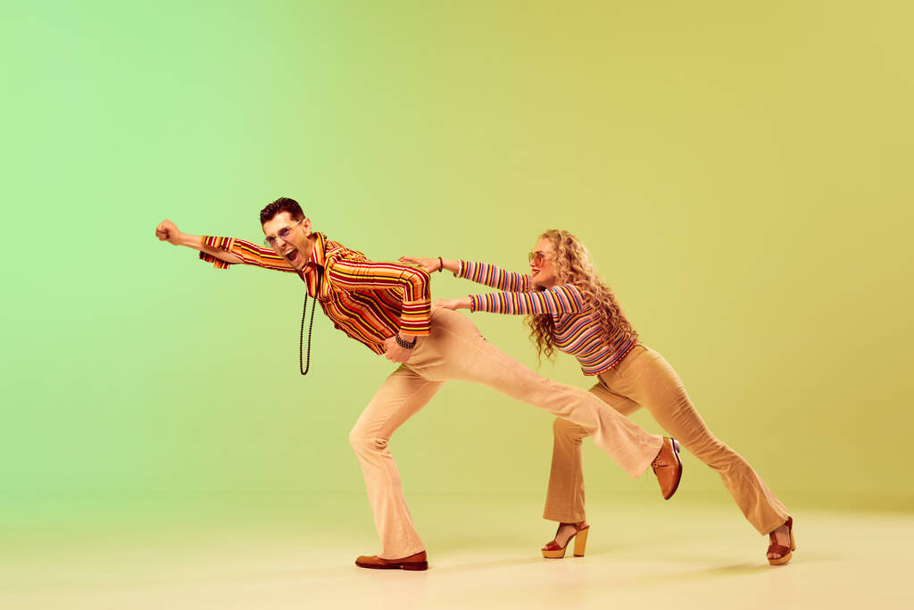 Pareja joven, expresiva y talentosa, hombre y mujer con elegantes trajes vintage bailando sobre un fondo verde degradado amarillo. Concepto de estilo retro, danza disco, moda, arte, hobby, música, años 70 - Foto, imagen