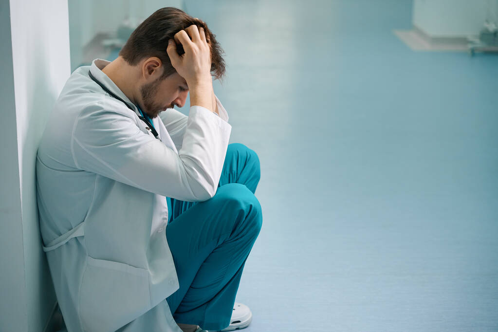 Triste joven interno en cuclillas en el pasillo del hospital, hombre en uniforme médico - Foto, imagen