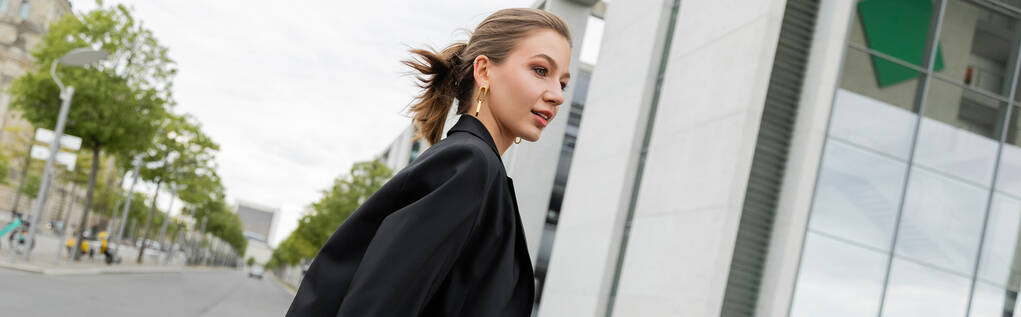 Mujer de pelo hermoso joven positivo en chaqueta negra mirando hacia otro lado mientras camina en Berlín, pancarta  - Foto, imagen