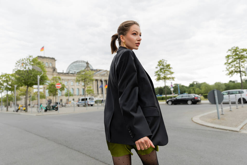 Fiatal, elegáns, szőke hajú nő fekete kabátban és ruhában, aki félrenéz, miközben Berlinben sétál - Fotó, kép