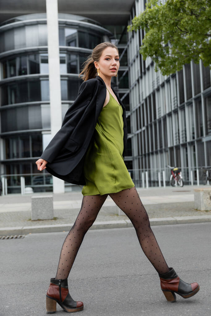 divatos fiatal nő zöld selyem ruhában, pöttyös harisnya és kabát nézi a kamerát Berlinben - Fotó, kép