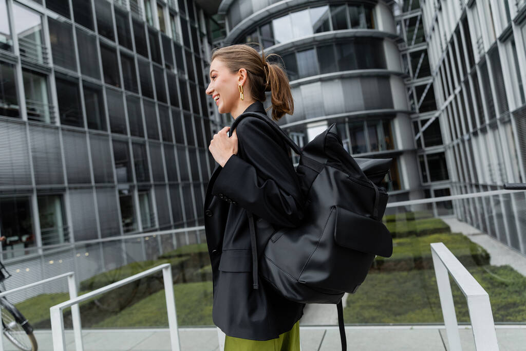Vue latérale de la jeune femme gaie et élégante en veste noire tenant le sac à dos et regardant loin  - Photo, image