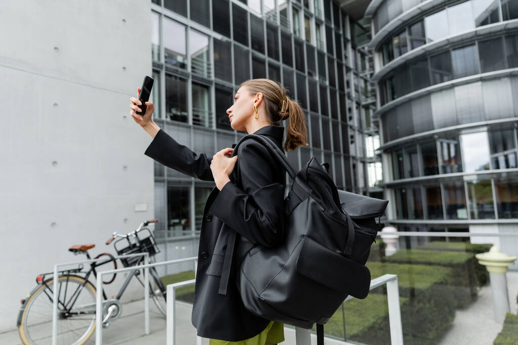 Seitenansicht der modischen und blonden jungen Frau mit Rucksack, die ein Selfie auf dem Smartphone macht - Foto, Bild