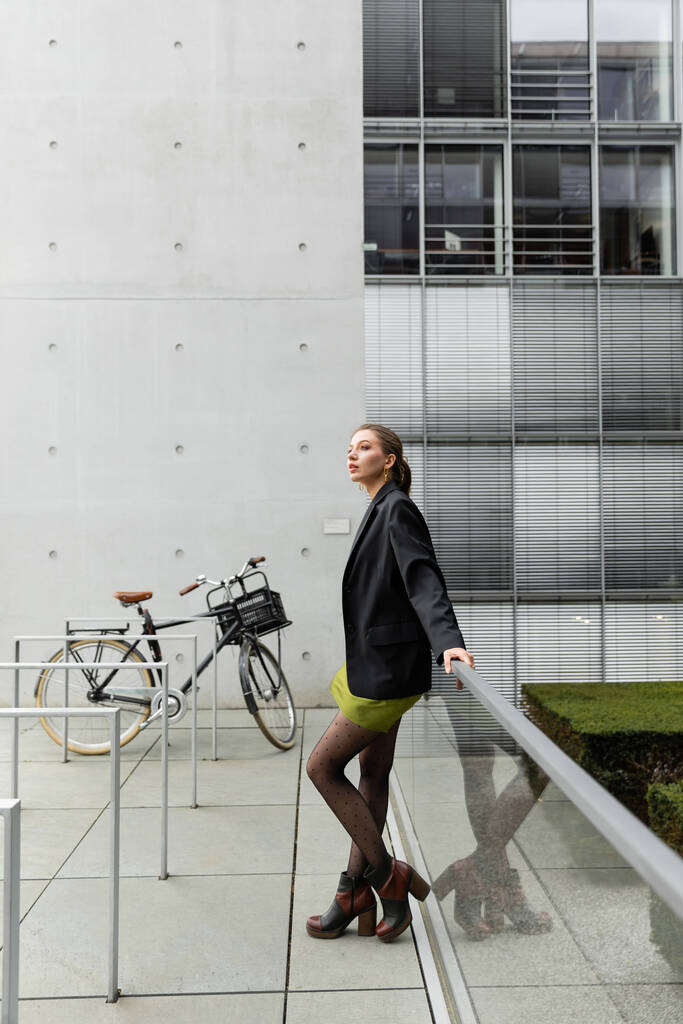 Mujer joven elegante y bonita en chaqueta negra y vestido de seda de pie cerca de la bicicleta en Berlín  - Foto, imagen