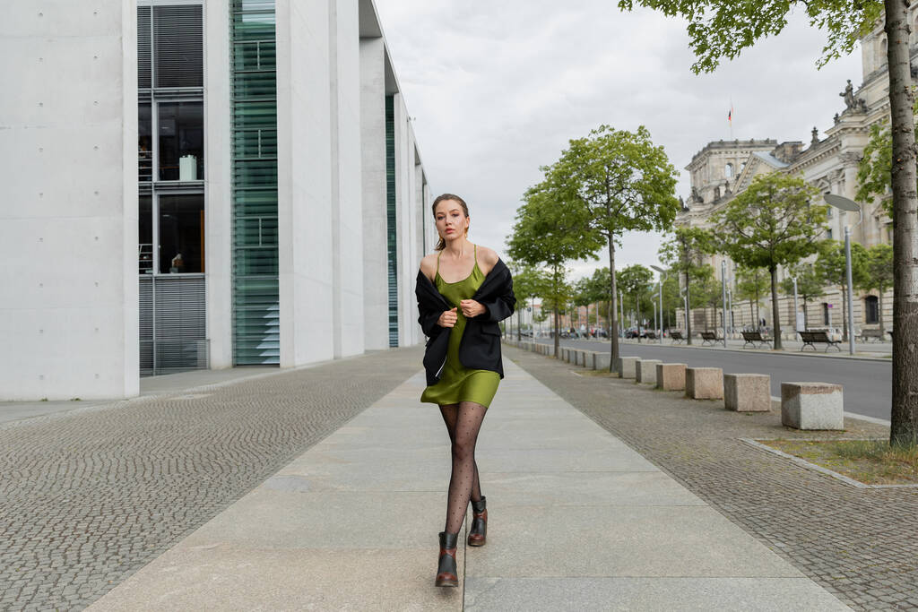 Berlin, Almanya 'da ceketli, ipek elbiseli ve botlu modaya uygun genç bir kadın kameraya bakıyor.  - Fotoğraf, Görsel