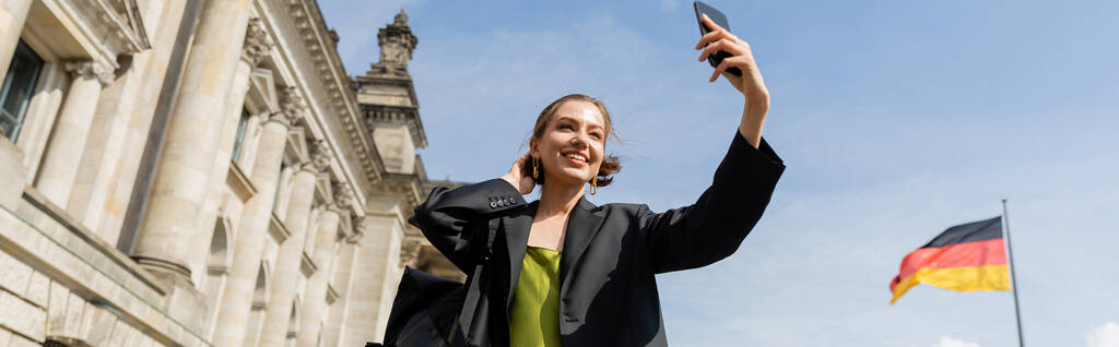 Mujer sonriente en blazer tomando selfie cerca del Reichstag Building en Berlín, pancarta - Foto, imagen