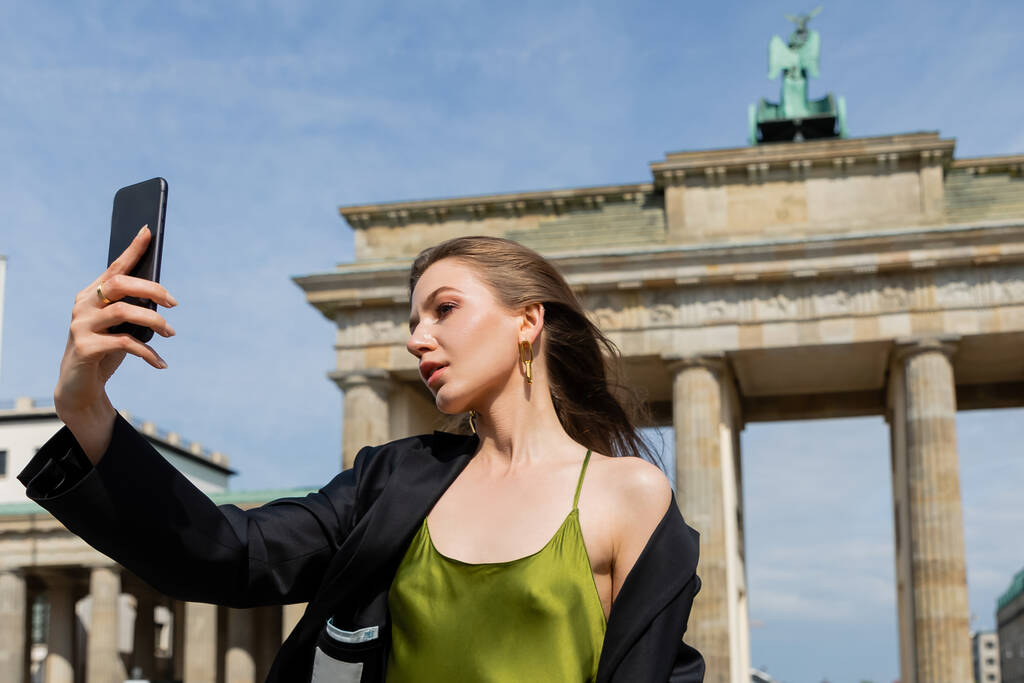 fiatal divatos nő dzsekiben és selyemruhában szelfit készít a berlini Brandenburg Gate közelében - Fotó, kép