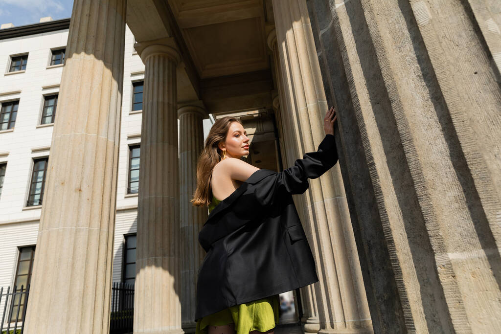 modna młoda kobieta w jedwabnej sukience trzymająca czarną kurtkę i stojąca w Berlinie, Niemcy  - Zdjęcie, obraz