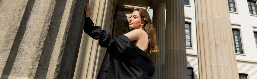 Elegant young woman in silk dress holding black jacket and standing in Berlin, banner - Photo, Image