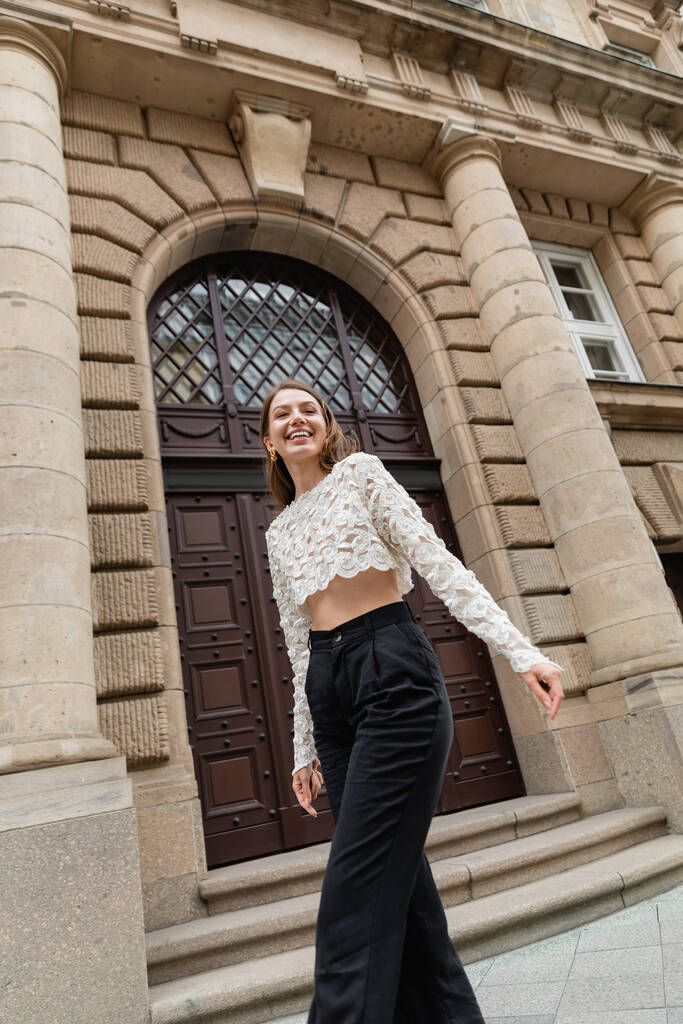 mujer joven optimista en top de encaje y pantalones de cintura alta mirando a la cámara en la calle urbana de Berlín - Foto, Imagen
