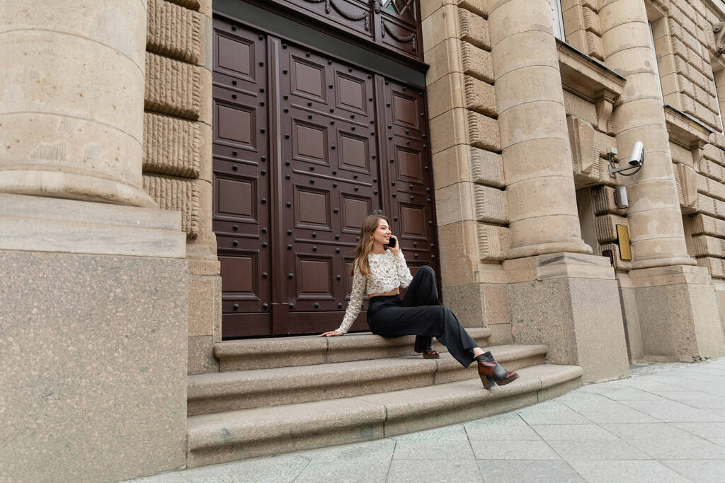 mujer sonriente hablando en el teléfono inteligente mientras está sentado en las escaleras cerca de la entrada del edificio en Berlín - Foto, Imagen