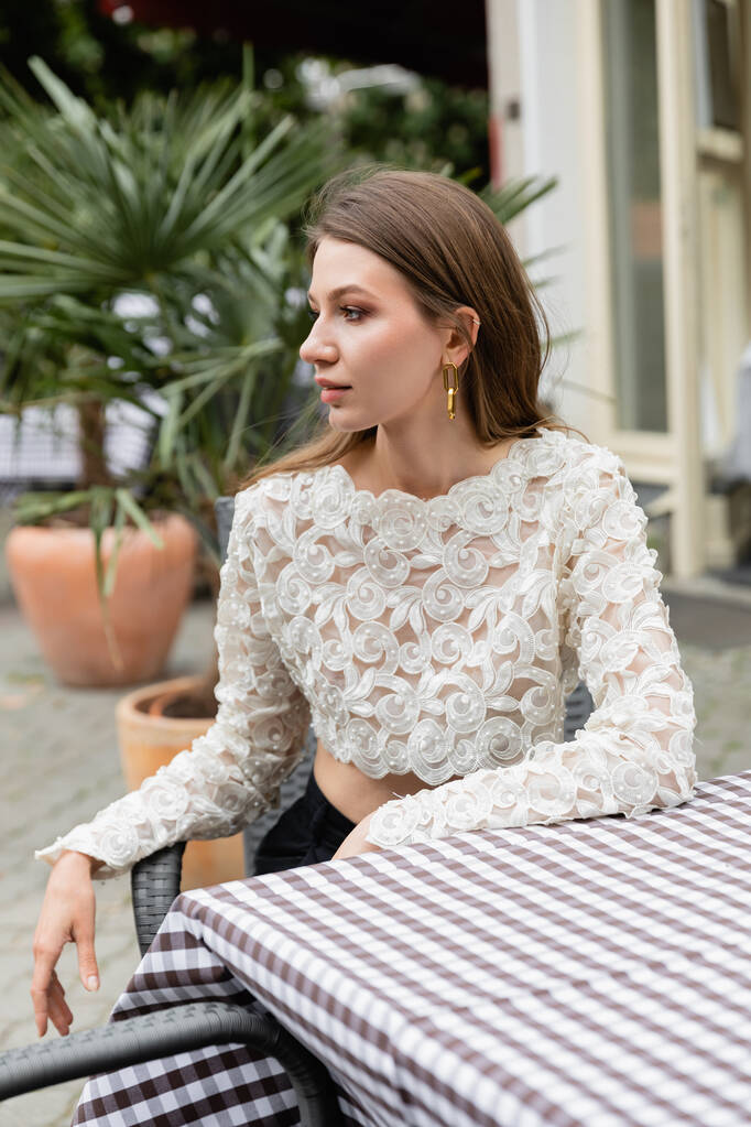 Mujer joven de moda en la parte superior de encaje blanco mirando hacia otro lado mientras está sentado cerca de la mesa en la terraza  - Foto, Imagen