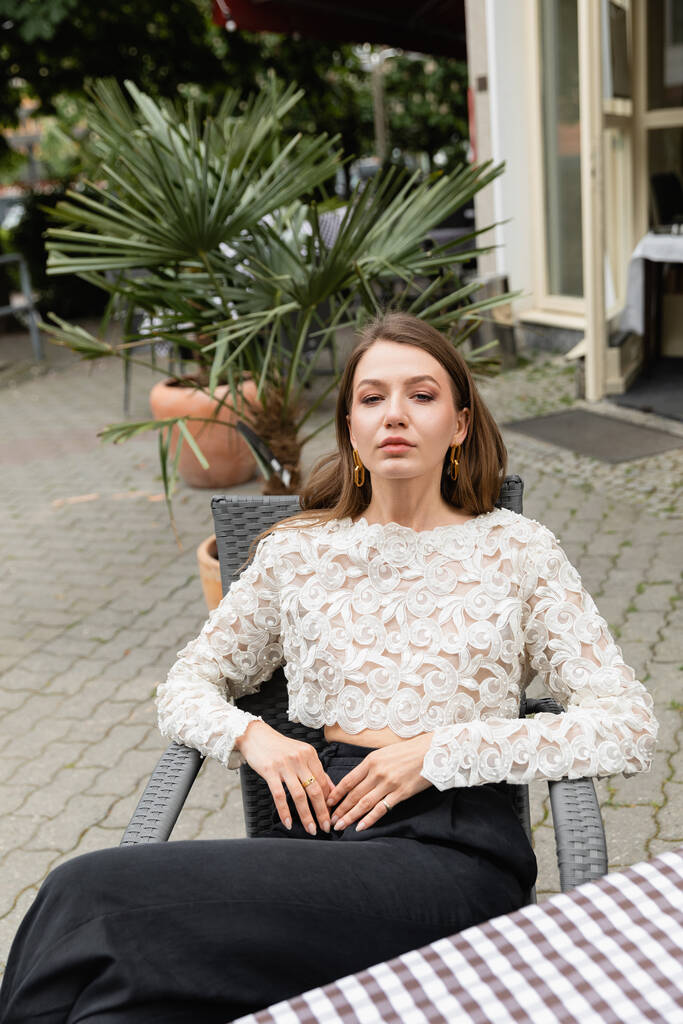 woman in stylish lace top and high waist pants looking at camera while sitting on terrace of cafe  - Photo, Image