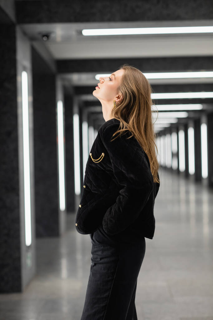 μοντέρνα νεαρή γυναίκα με μαύρο μπουφάν ποζάρει σε μοντέρνο κτίριο με λαμπτήρες φθορισμού στο Βερολίνο - Φωτογραφία, εικόνα