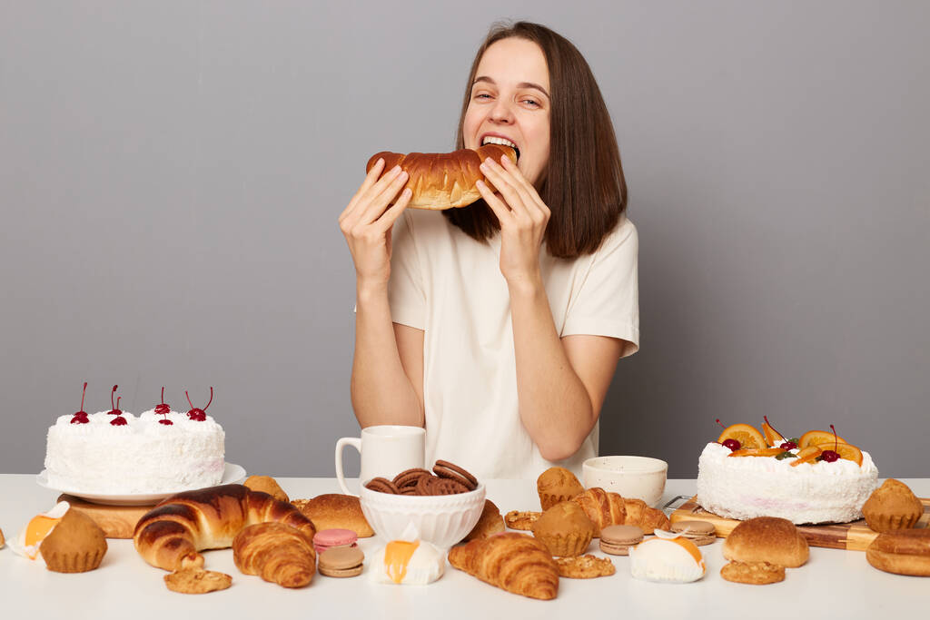 Comida chatarra, culinaria, panadería. Mujer divertida hambrienta vistiendo camiseta blanca comiendo con buen apetito bagel posa en la mesa festiva con varios postres, aislado sobre fondo gris. - Foto, Imagen