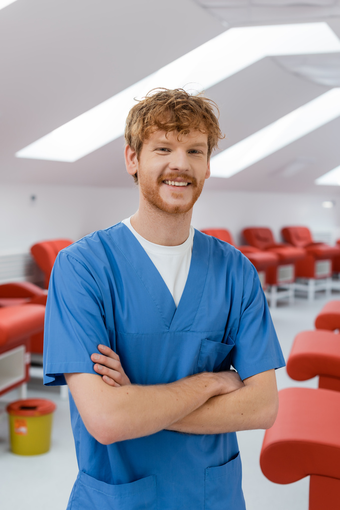 κοκκινομάλλα υπάλληλος υγείας με μπλε στολή στέκεται με διπλωμένα χέρια και χαμογελώντας στην κάμερα κοντά σε άνετες ιατρικές καρέκλες στο κέντρο αιμοδοσίας, θολή φόντο - Φωτογραφία, εικόνα