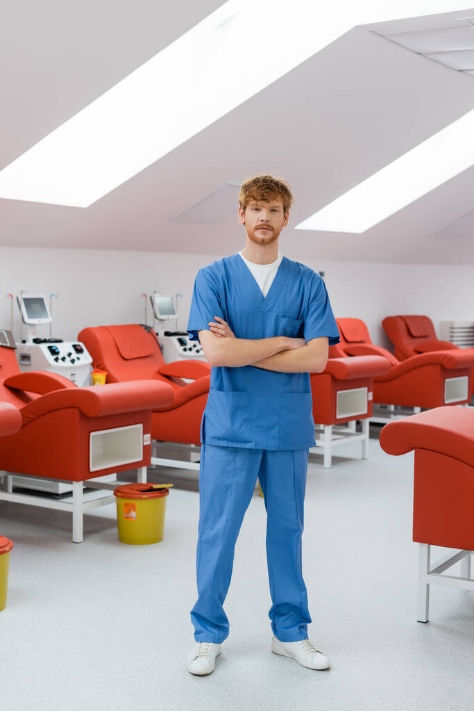 plná délka zrzky a mladého lékaře v modré uniformě stojící se založenýma rukama v blízkosti pohodlných lékařských židlí, automatizovaných transfuzních strojů a koše na odpadky v dárcovském centru - Fotografie, Obrázek