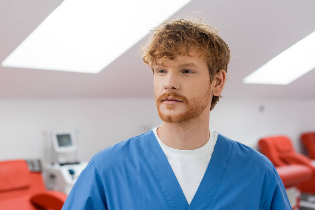 молодой, рыжий и бородатый доктор в синей униформе стоит и смотрит в сторону размытых медицинских стульев в стерильной среде станции переливания крови - Фото, изображение