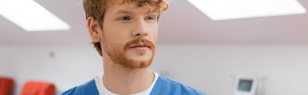 young, redhead healthcare worker in blue uniform looking away in sterile environment of laboratory in blood donation center on blurred background, banner - Photo, Image