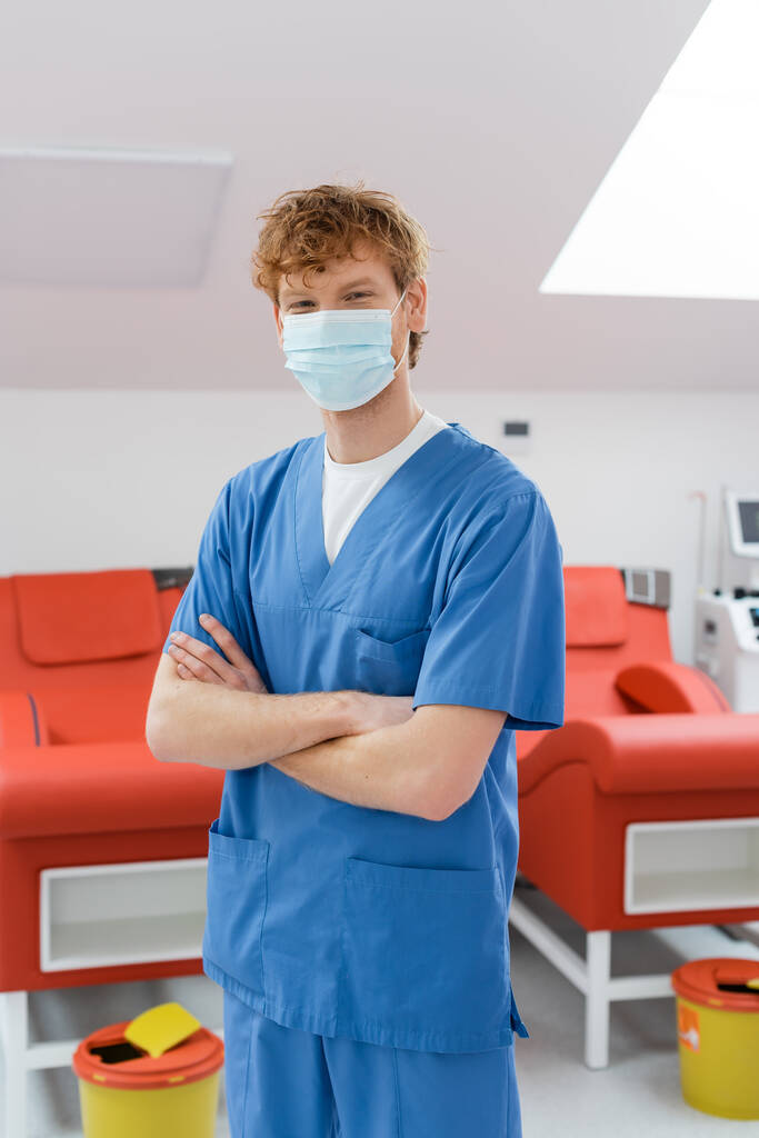 Junge rothaarige Ärztin in medizinischer Maske und blauer Uniform steht mit verschränkten Armen und blickt in die Kamera in der Nähe von Medizinstühlen mit ergonomischem Design und Mülleimern im Blutspendezentrum - Foto, Bild