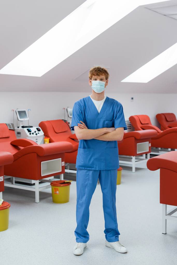 Mavi üniformalı ve tıbbi maskeli kızıl saçlı bir doktor modern hastanedeki tıbbi koltukların, nakil makinelerinin ve çöp kovalarının yanında duruyor. - Fotoğraf, Görsel