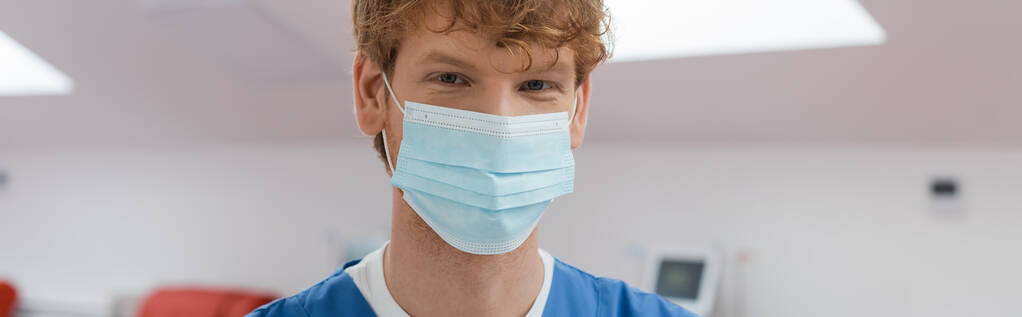 mladý a zrzavý lékař s veselým výrazem v očích, nosí lékařskou masku a dívá se do kamery ve sterilním prostředí centra dárcovství krve na rozmazaném pozadí, banner - Fotografie, Obrázek