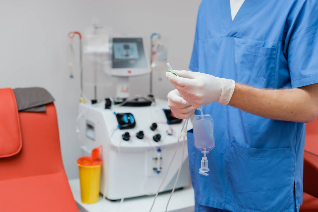 青い制服と滅菌ラテックス手袋の医師のクロップドビューは、研究室でぼやけた自動機器やプラスチックカップの近くに血の輸血セットを保持 - 写真・画像