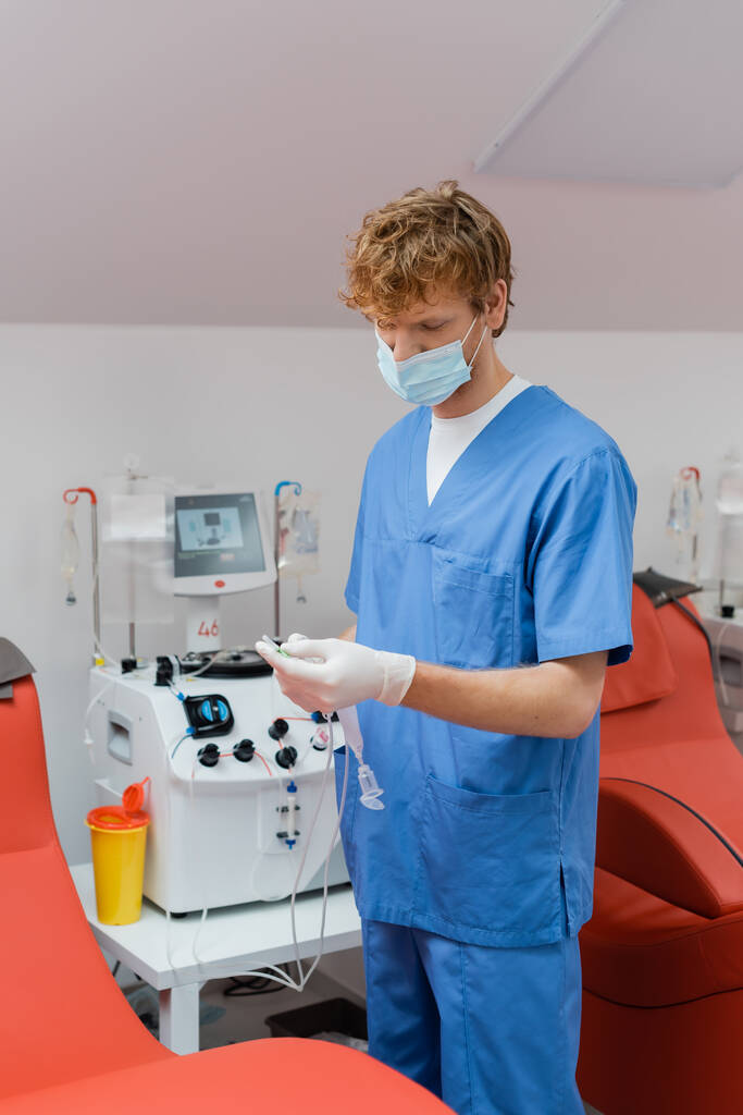 Rotschopf und junger Arzt in medizinischer Maske, Latex-Handschuhen und blauer Uniform halten Transfusionsset in der Nähe von automatisierten Geräten und medizinischen Stühlen im Blutspendezentrum - Foto, Bild