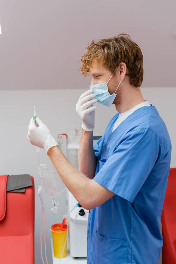oldalnézet fiatal és vörös hajú orvos kék egyenruhában, orvosi maszk és latex kesztyű tartja vérátömlesztés készlet közelében orvosi székek steril laboratóriumban - Fotó, kép
