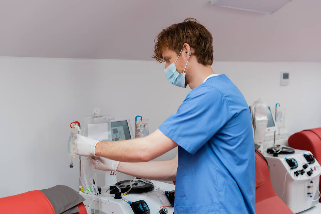 travailleur de la santé roux en masque médical, uniforme bleu et gants en latex tenant ensemble de transfusion près du stand de goutte à goutte, chaises médicales et équipement automatisé en laboratoire - Photo, image