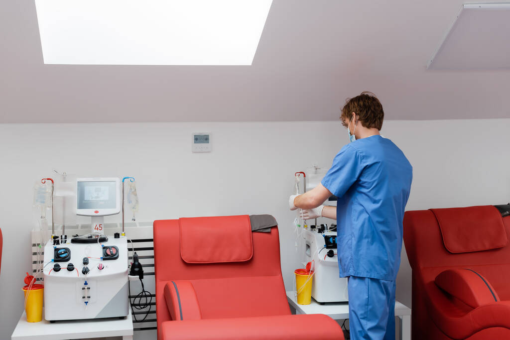 πίσω όψη της νεαρής κοκκινομάλλας γιατρός με μπλε στολή κοντά σε αυτοματοποιημένα μηχανήματα μετάγγισης, πλαστικά ποτήρια, βάσεις για σταγόνες και άνετες ιατρικές καρέκλες στο κέντρο αιμοδοσίας - Φωτογραφία, εικόνα