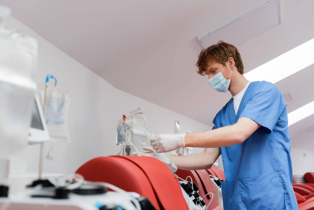 zrzka zdravotnický pracovník v lékařské masce a latexové rukavice montáž transfuzní set v blízkosti automatizovaného vybavení a lékařské židle v centru darování krve - Fotografie, Obrázek