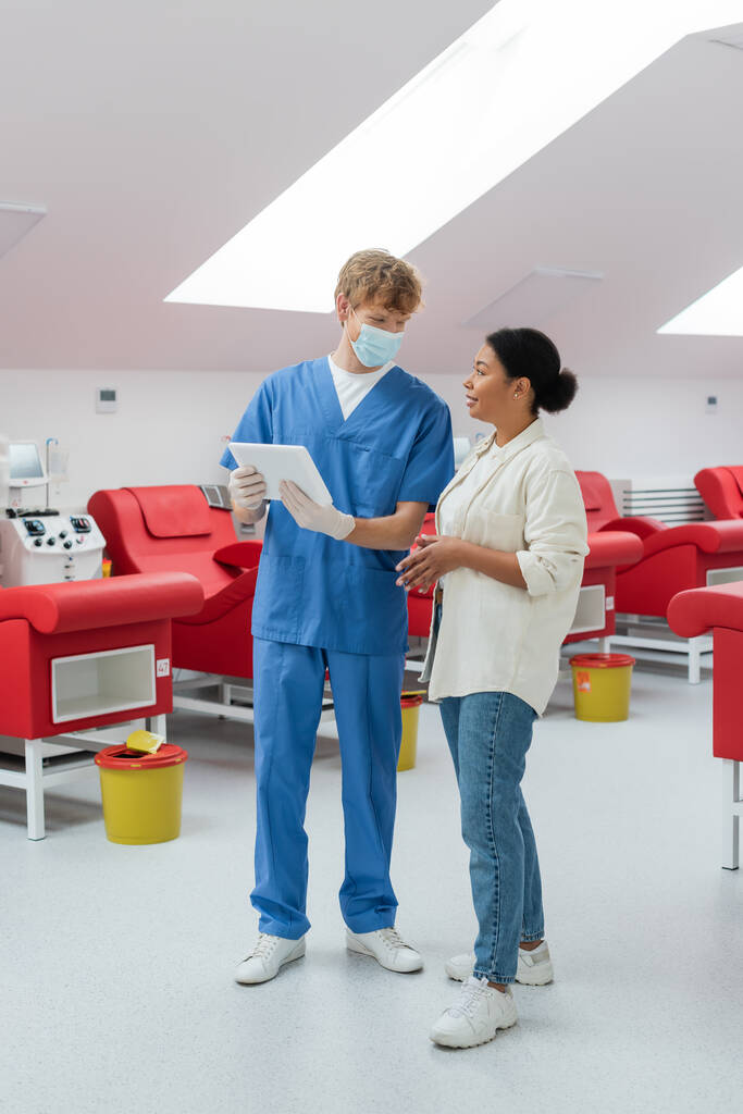 redhead doctor in blue uniform, medical mask and latex gloves holding digital tablet and talking to multiracial woman near medical chairs, transfusion machine and trash buckets in laboratory - Photo, Image