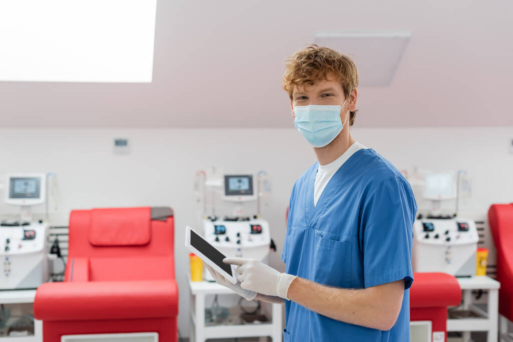 trabajador sanitario pelirrojo en uniforme azul, máscara médica y guantes de látex mirando a la cámara cerca de sillas médicas y máquinas de transfusión sobre fondo borroso en el centro de donación de sangre - Foto, Imagen
