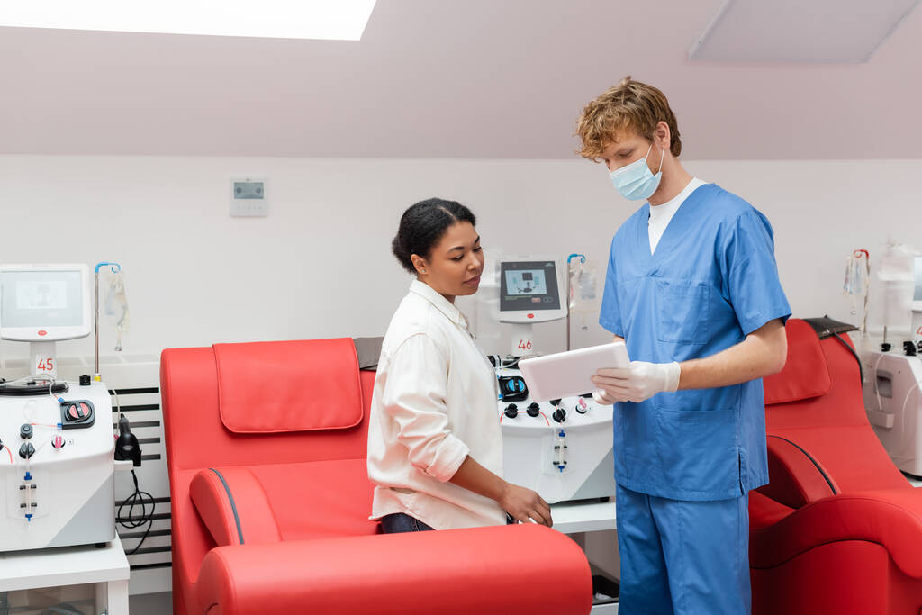 Arzt in medizinischer Maske, blauer Uniform und Latex-Handschuhen zeigt der multiethnischen Frau, die auf einem bequemen ergonomischen Stuhl in der Nähe von Transfusionsgeräten im Blutspendezentrum sitzt, ein digitales Tablet - Foto, Bild