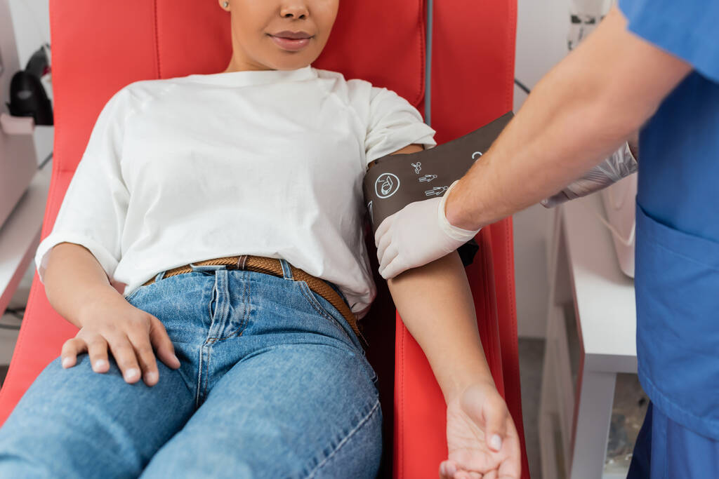 частковий вигляд лікаря в латексних рукавичках, що регулює тиск манжети на багаторасову жінку, що сидить на зручному медичному кріслі в станції переливання крові, догляд за пацієнтами
 - Фото, зображення