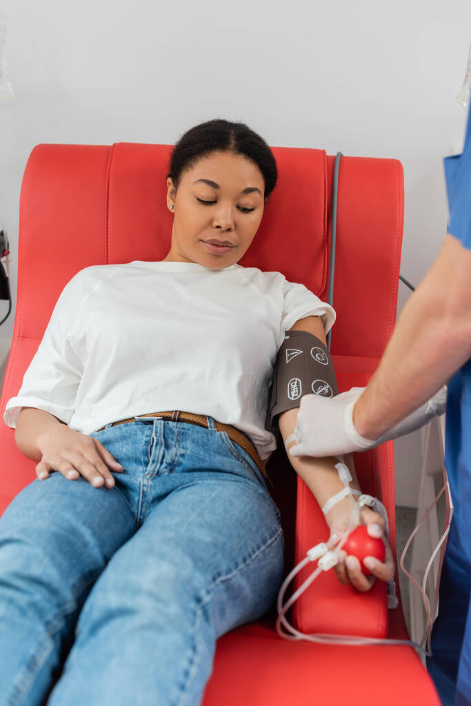 travailleur de la santé reliant la transfusion sanguine à une femme multiraciale assise sur une chaise médicale confortable dans un brassard de pression artérielle et serrant une balle en caoutchouc - Photo, image