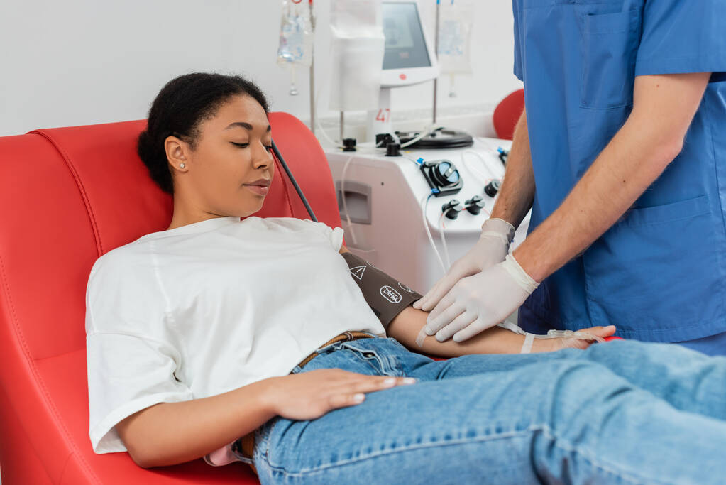 medico in guanti di lattice attaccare cerotto sul braccio della donna multirazziale seduto sulla sedia medica e donare il sangue vicino alla macchina trasfusione automatizzata - Foto, immagini