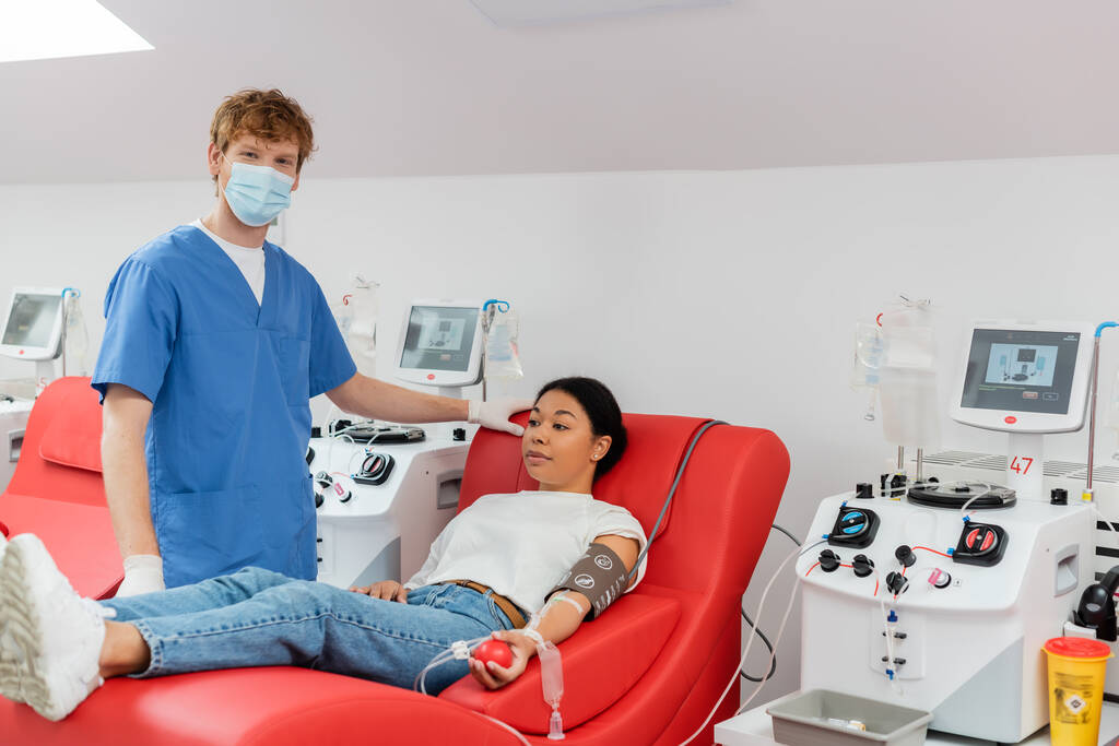γιατρός με ιατρική μάσκα, μπλε στολή και γάντια λατέξ κοιτάζοντας κάμερα κοντά σε μηχανές μετάγγισης και πολυφυλετική γυναίκα κάθεται σε ιατρική καρέκλα, ενώ αιμοδοσία στην κλινική - Φωτογραφία, εικόνα