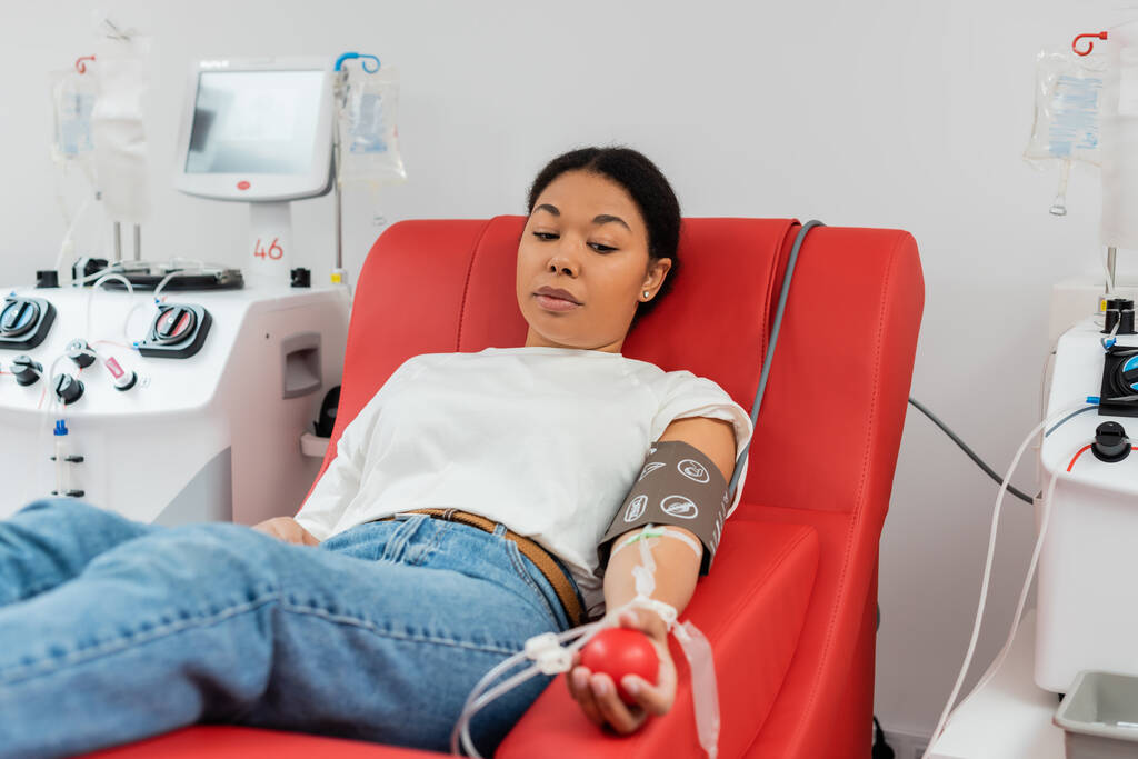 многорасовая женщина с набором для переливания крови и резиновым мячом сидит на эргономичном медицинском стуле рядом с аппаратом для переливания крови и сдает кровь в больнице - Фото, изображение